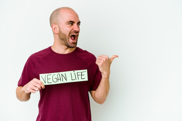 Jovem homem careca caucasiano segurando um cartaz de vida Vegan isolado em pontos de fundo roxo com o dedo polegar afastado, rindo e despreocupado.