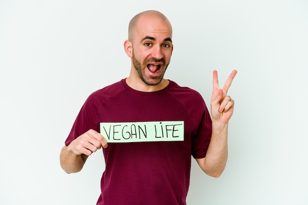 Jovem homem careca caucasiano segurando um cartaz de vida Vegan isolado no fundo roxo, alegre e despreocupado, mostrando um símbolo de paz com os dedos.