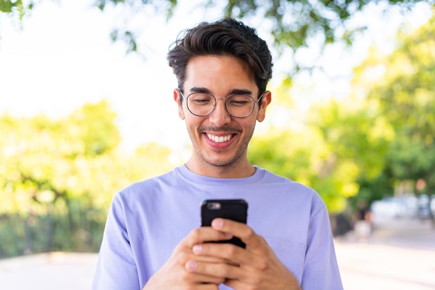 Jovem homem caucasiano ao ar livre em um parque enviando uma mensagem com o celular