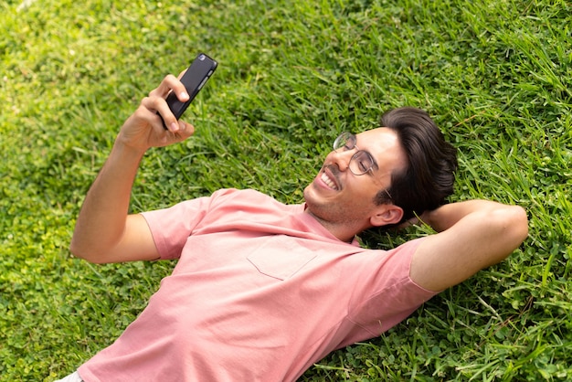 Jovem homem caucasiano ao ar livre em um parque usando telefone celular com expressão feliz