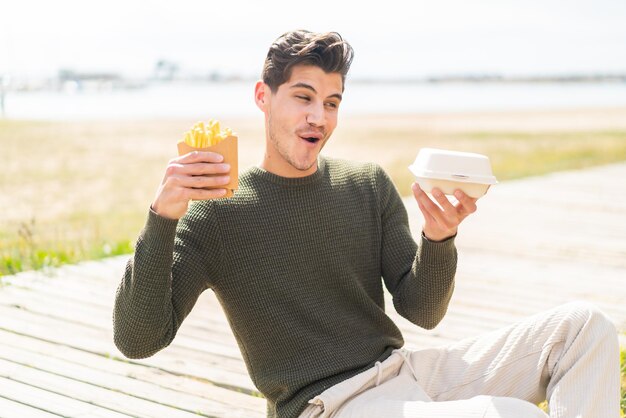 Jovem homem caucasiano ao ar livre levando batatas fritas e comida de caixa para viagem com expressão de surpresa