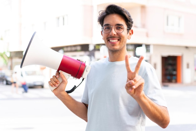 Jovem homem caucasiano ao ar livre segurando um megafone e sorrindo e mostrando sinal de vitória