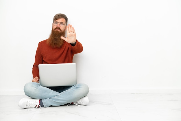 Jovem homem caucasiano avermelhado com laptop isolado em fundo branco fazendo gesto de parada