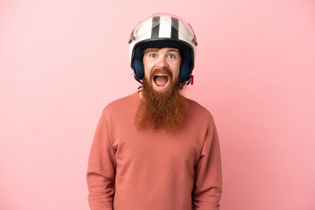 Jovem homem caucasiano avermelhado com um capacete de moto isolado em fundo rosa com expressão facial de surpresa