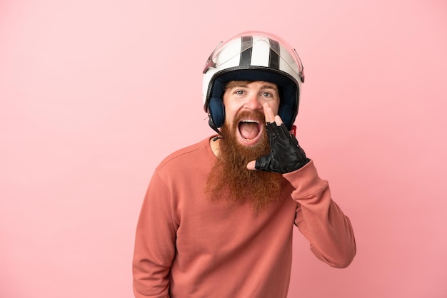 Jovem homem caucasiano avermelhado com um capacete de moto isolado em fundo rosa gritando com a boca aberta