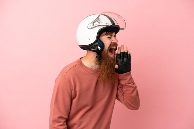 Jovem homem caucasiano avermelhado com um capacete de moto isolado no fundo rosa gritando com a boca aberta para o lado