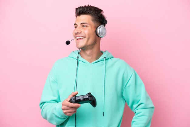 Jovem homem caucasiano bonito brincando com um controlador de videogame isolado em fundo rosa posando com braços no quadril e sorrindo