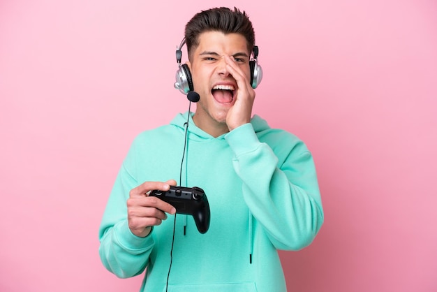Jovem homem caucasiano bonito brincando com um controlador de videogame isolado no fundo rosa gritando com a boca aberta