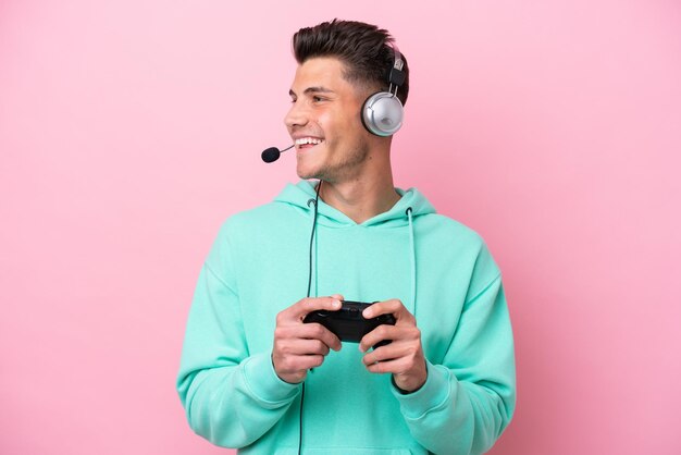 Jovem homem caucasiano bonito brincando com um controlador de videogame isolado no fundo rosa, olhando para o lado e sorrindo