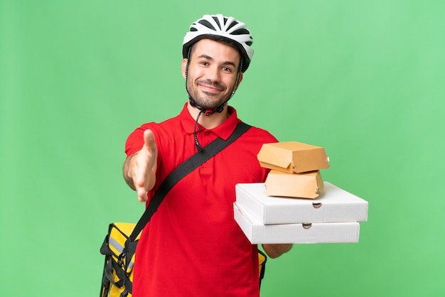 Jovem homem caucasiano bonito com mochila térmica e segurando comida para viagem sobre fundo isolado, apertando as mãos para fechar um bom negócio