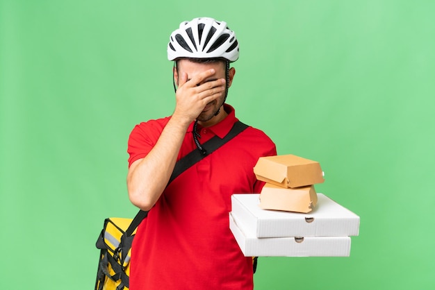 Jovem homem caucasiano bonito com mochila térmica e segurando comida para viagem sobre fundo isolado com expressão cansada e doente