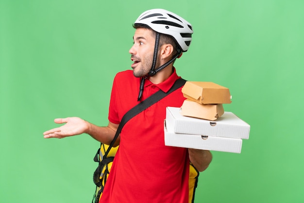 Foto jovem homem caucasiano bonito com mochila térmica e segurando comida para viagem sobre fundo isolado com expressão de surpresa enquanto olha de lado