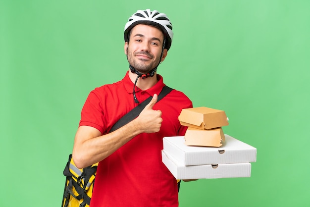 Jovem homem caucasiano bonito com mochila térmica e segurando comida para viagem sobre fundo isolado, dando um polegar para cima gesto