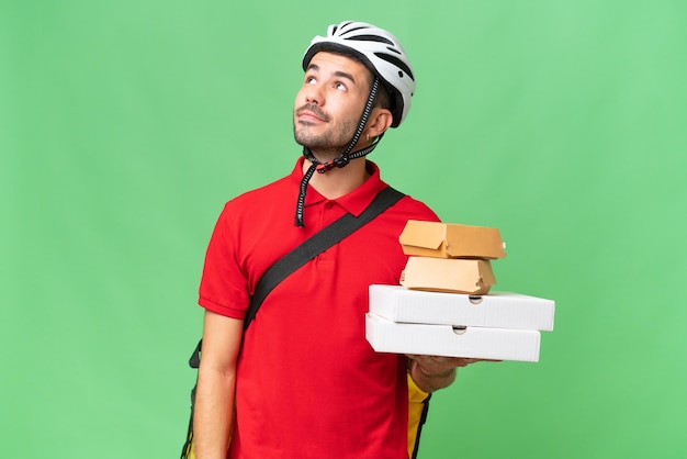 Foto jovem homem caucasiano bonito com mochila térmica e segurando comida para viagem sobre fundo isolado, pensando em uma ideia enquanto olha para cima