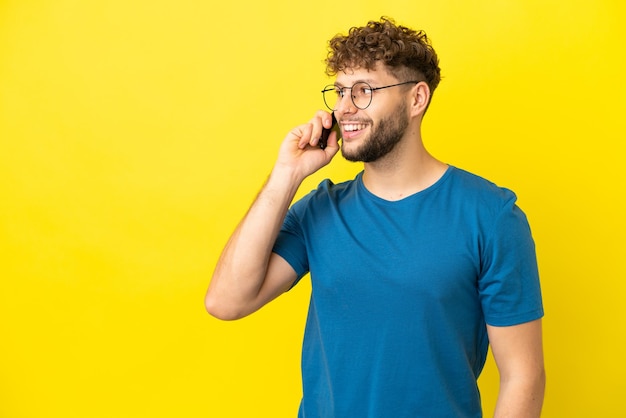 Jovem homem caucasiano bonito isolado em fundo amarelo, mantendo uma conversa com o telefone celular