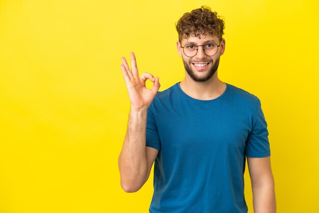 Jovem homem caucasiano bonito isolado em fundo amarelo mostrando sinal de ok com os dedos