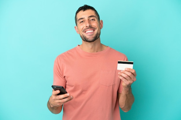 Jovem homem caucasiano bonito isolado em fundo azul comprando com o celular com cartão de crédito