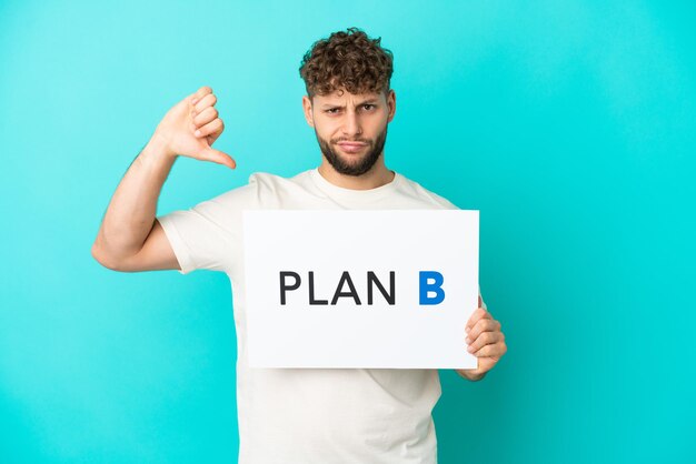 Jovem homem caucasiano bonito isolado em fundo azul segurando um cartaz com a mensagem PLANO B e fazendo sinal ruim