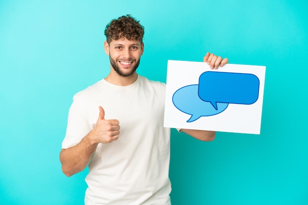 Jovem homem caucasiano bonito isolado em fundo azul segurando um cartaz com ícone de bolha do discurso com o polegar para cima