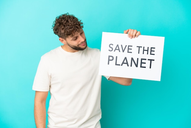 Jovem homem caucasiano bonito isolado em fundo azul segurando um cartaz com texto Salve o Planeta