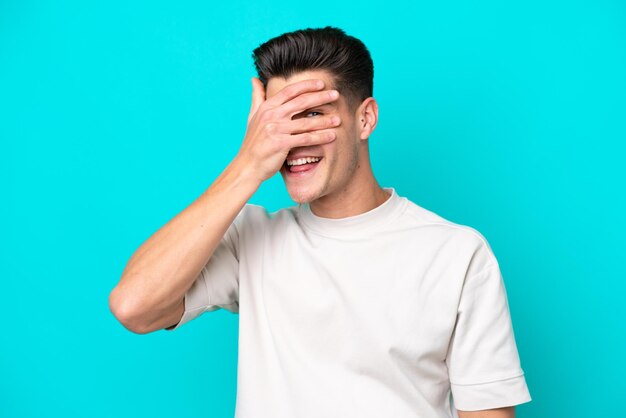 Jovem homem caucasiano bonito isolado no fundo azul cobrindo os olhos pelas mãos e sorrindo