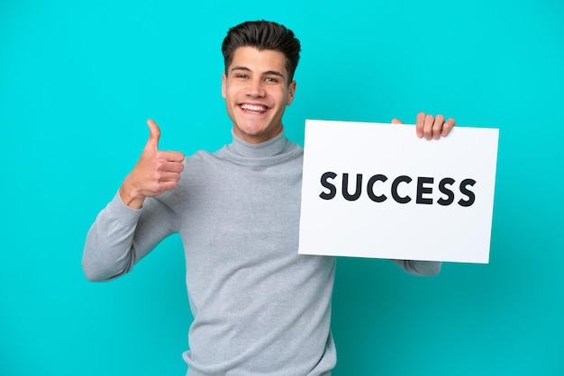 Jovem homem caucasiano bonito isolado no fundo azul segurando um cartaz com texto SUCESSO com o polegar para cima