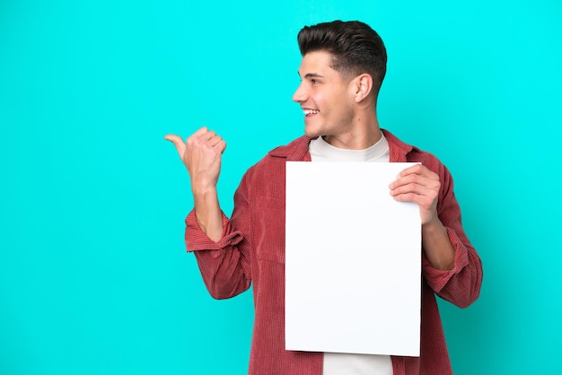 Foto jovem homem caucasiano bonito isolado no fundo azul segurando um cartaz vazio e apontando o lado