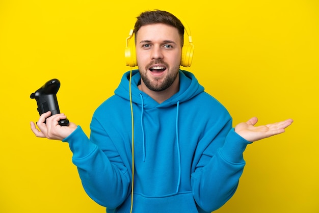 Jovem homem caucasiano brincando com um controlador de videogame isolado em fundo amarelo com expressão facial chocada