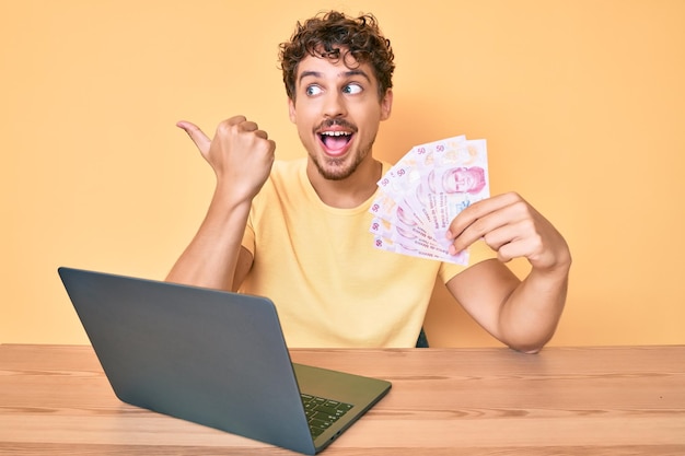 Jovem homem caucasiano com cabelo encaracolado sentado na mesa trabalhando com laptop e segurando notas de pesos mexicanos apontando o polegar para o lado sorrindo feliz com a boca aberta