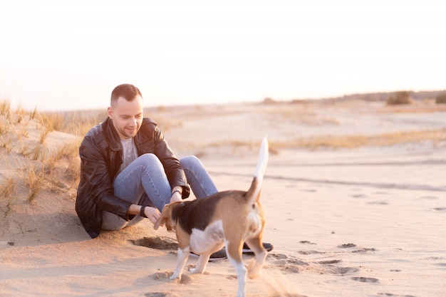 Jovem homem caucasiano com cachorro Beagle senta-se na praia