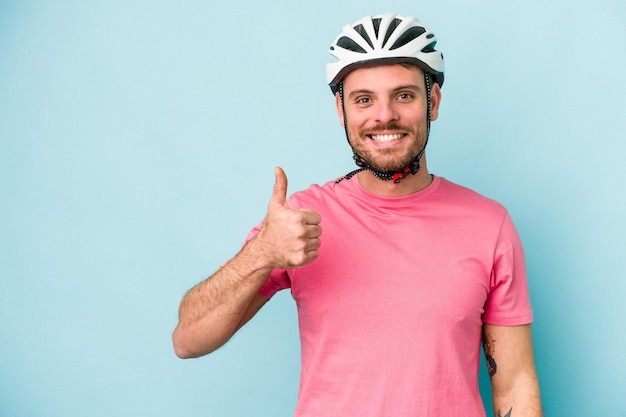 Jovem homem caucasiano com capacete de bicicleta isolado em fundo azul sorrindo e levantando o polegar