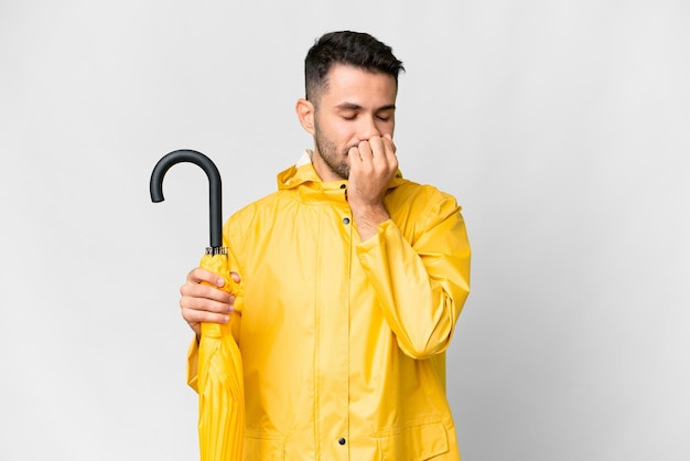 Jovem homem caucasiano com casaco à prova de chuva e guarda-chuva sobre fundo branco isolado com dúvidas