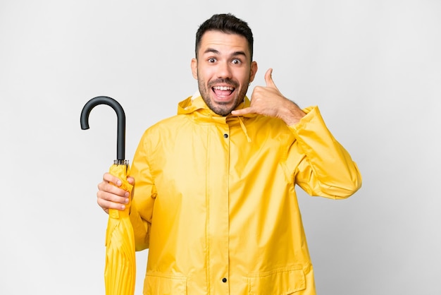 Jovem homem caucasiano com casaco à prova de chuva e guarda-chuva sobre fundo branco isolado, fazendo gesto de telefone Ligue-me de volta sinal