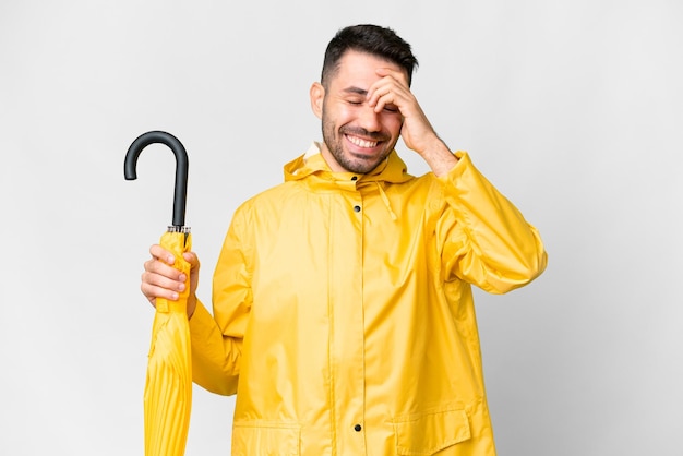 Jovem homem caucasiano com casaco à prova de chuva e guarda-chuva sobre fundo branco isolado rindo