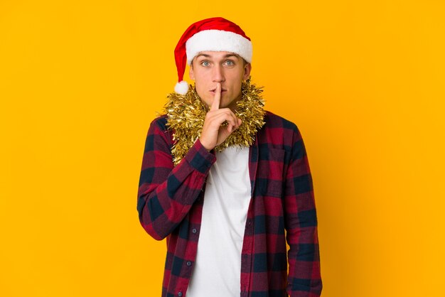Jovem homem caucasiano com chapéu de Natal segurando um presente isolado em um fundo amarelo, mantendo um segredo ou pedindo silêncio.