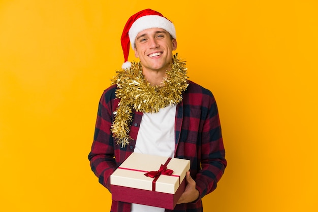 Jovem homem caucasiano com chapéu de Natal segurando um presente isolado em um fundo amarelo se sente orgulhoso e autoconfiante, exemplo a seguir.