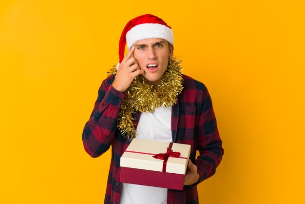 Jovem homem caucasiano com chapéu de Natal segurando um presente isolado em um fundo amarelo, sendo chocado, ela se lembrou de uma reunião importante.