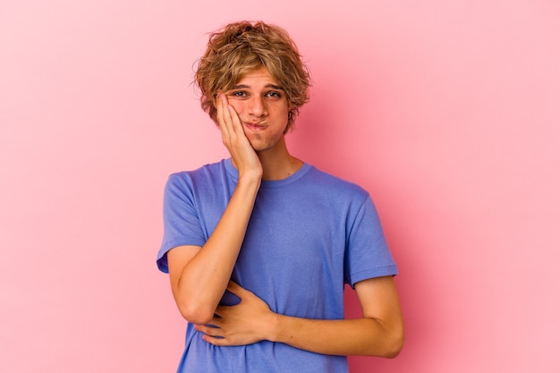 Jovem homem caucasiano com maquiagem isolada no fundo rosa sopra nas bochechas, tem expressão de cansaço. Conceito de expressão facial.