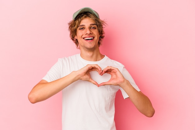 Jovem homem caucasiano com maquiagem isolada no fundo rosa, sorrindo e mostrando uma forma de coração com as mãos.