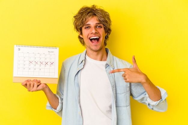 Jovem homem caucasiano com maquiagem segurando calendário isolado em fundo amarelo pessoa apontando com a mão para um espaço de cópia de camisa, orgulhoso e confiante