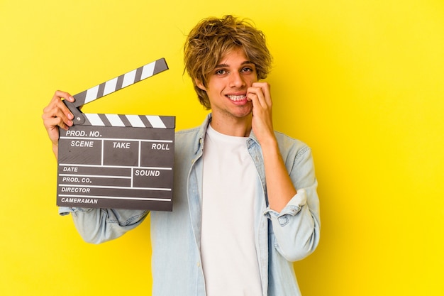 Jovem homem caucasiano com maquiagem segurando claquete isolada em fundo amarelo, roendo as unhas, nervoso e muito ansioso.