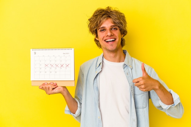 Jovem homem caucasiano com maquiagem segurando o calendário isolado no fundo amarelo, sorrindo e levantando o polegar
