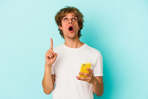 Jovem homem caucasiano com maquiagem segurando o celular isolado em um fundo azul apontando para cima com a boca aberta.