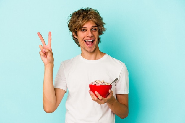 Jovem homem caucasiano com maquiagem uma tigela de cereal isolada no fundo azul, alegre e despreocupada, mostrando um símbolo de paz com os dedos.