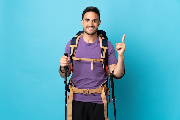 Jovem homem caucasiano com mochila e bastões de trekking isolados em azul apontando para uma ótima ideia