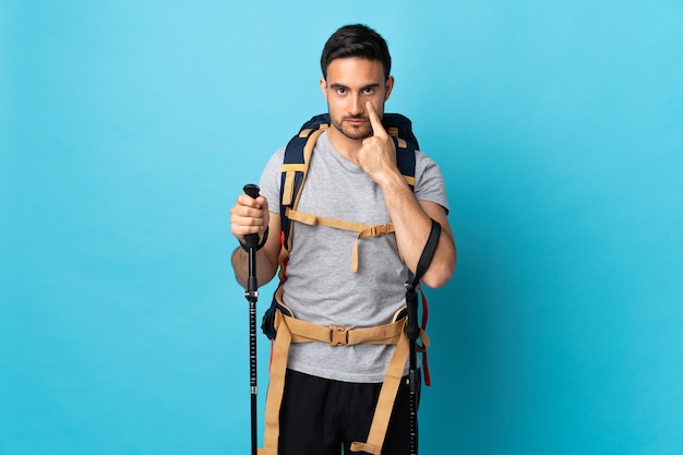 Jovem homem caucasiano com mochila e bastões de trekking isolados na parede azul mostrando algo