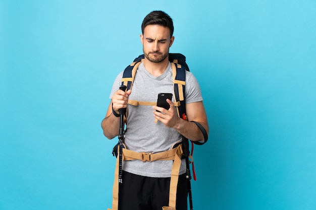 Jovem homem caucasiano com mochila e bastões de trekking isolados no azul, pensando e enviando uma mensagem