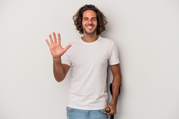 Jovem homem caucasiano com muletas isoladas em fundo cinza, sorrindo alegre mostrando o número cinco com os dedos.