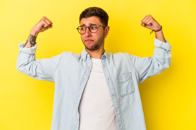 Jovem homem caucasiano com tatuagens isoladas em fundo amarelo, mostrando força gesto com os braços, símbolo do poder feminino