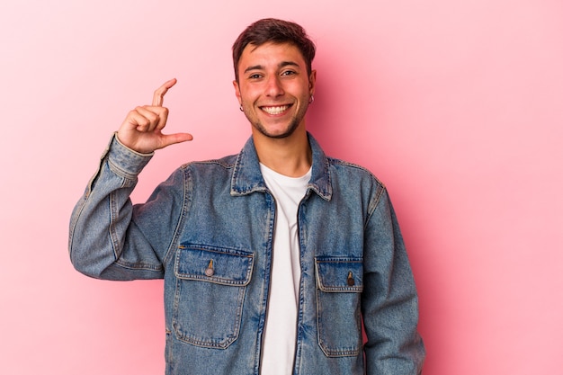 Jovem homem caucasiano com tatuagens isoladas em fundo amarelo, segurando algo pequeno com os indicadores, sorrindo e confiante.